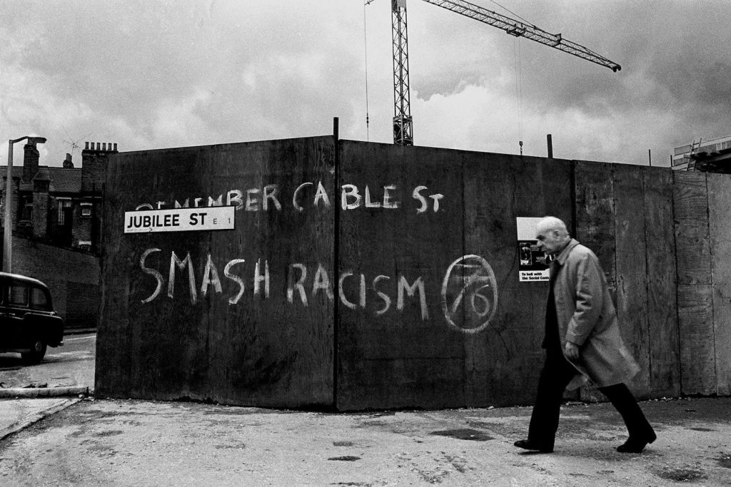 A man walking past an industrial site, with a graffiti commemorating the Battle of Cable Street, Stepney, 1977, Whitechapel.