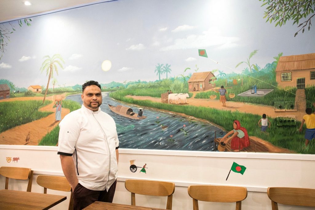 Chef Atik posing beside a painting of a Bangladeshi village, Brick Lane.