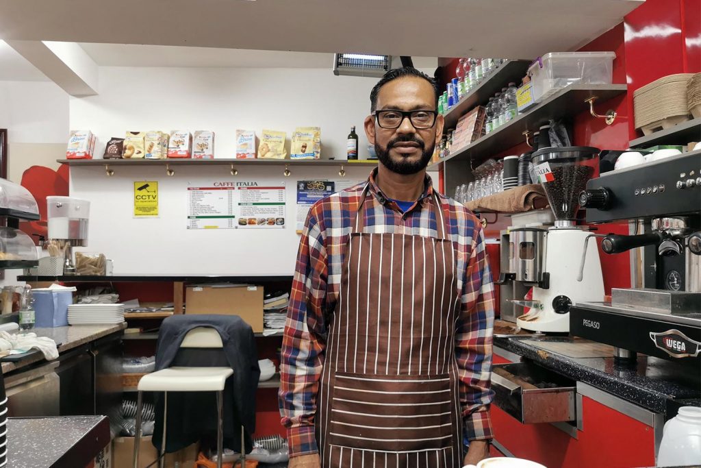 A man in an apron standing behind the counter of a cafe with a big coffee machine next to him, Whitechapel, London
